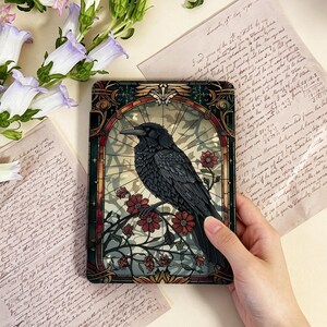 Pode incluir: Uma capa para tablet com um design de corvo preto. A obra de arte apresenta um corvo empoleirado num galho com flores vermelhas, contra um fundo de vitral. A capa é segurada por uma mão, com elementos florais e papel vintage no fundo.