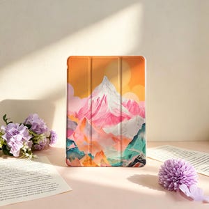Op de afbeelding: Een rechthoekige tablethoes met een levendige grafische print van een besneeuwd gebergte tegen een oranje en gele zonsonderganghemel. Het ontwerp bevat tinten roze, wit, blauwgroen en oranje, met een zichtbare drievoudig opvouwbare hoes.
