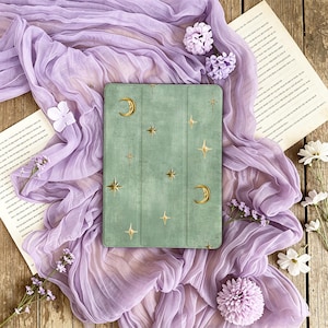 May include: A sage green tablet case featuring a gold celestial pattern of crescent moons and four-pointed stars. The rectangular cover has a textured finish and a tri-fold design, displaying a consistent motif of golden stars across the surface.