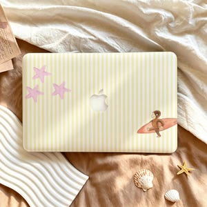May include: A laptop cover featuring a light yellow and white vertical striped pattern. The design includes three light pink starfish on the left side and a small illustration of a brown monkey carrying a surfboard on the right side.