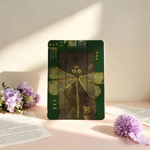 May include: A rectangular tablet case featuring a dark green background with a central dark brown vertical panel. A gold-colored dragonfly illustration is centered on the case, accompanied by small gold geometric accents and two small square icons.