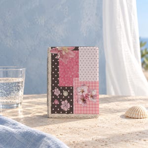 May include: A rectangular e-reader case featuring a patchwork design of pink and brown panels. The pattern includes polka dots, plaid, and floral motifs with small decorative buttons. The edges are trimmed with a lace-like border in a neutral tone.