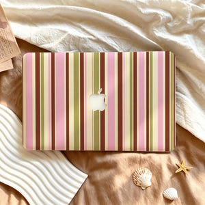 Può includere: Una cover per laptop con un motivo a righe verticali in tonalità alternate di rosa chiaro, verde oliva, crema e marrone scuro. Il design presenta un ritaglio centrale che rivela il logo Apple argentato sulla superficie del laptop.