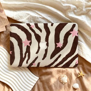Op de afbeelding: Een harde laptophoes met een bruin-wit zebrapatroon en diverse lichtroze stervormige stickers verspreid over het oppervlak. Het midden van de hoes heeft een uitsparing voor het Apple-logo, waardoor de zilverkleurige afwerking zichtbaar is.