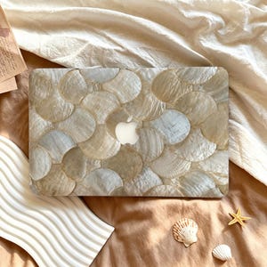 May include: A laptop cover featuring a repeating pattern of overlapping, iridescent, cream and beige shell-like circular tiles. The surface has a pearlescent, shimmering finish with a central cutout revealing the silver Apple logo on the device.