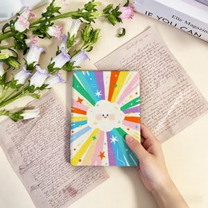 May include: A colorful notebook with a cloud and rainbow design. The cover features a smiling cloud at the center, surrounded by radiating stripes in various colors. The notebook is held by a hand, with flowers and vintage papers in the background.