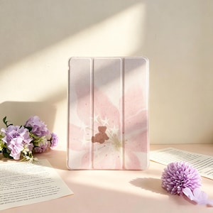 May include: A rectangular tablet case featuring a soft pink floral pattern with a small brown teddy bear illustration and white star accents. The case has a tri-fold design with vertical creases and a smooth, matte finish on the front cover.