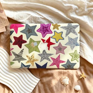 May include: A laptop skin featuring a cream-colored background covered in a variety of star shapes. The stars display diverse textures and patterns, including bright pink, yellow, silver, blue, green, and zebra-striped designs, with a central cutout.