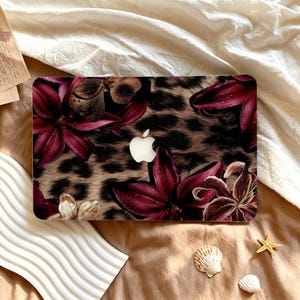 Könnte beinhalten: Eine Laptop-Hülle mit einem braun-schwarzen Leopardenmuster, das mit dunkelroten Lilien überlagert ist. Das Design zeigt Wassertropfen auf den Blütenblättern und einen mittigen Ausschnitt, der das weiße Apple-Logo auf dem Deckel freigibt.