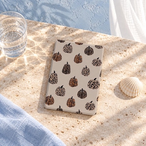 May include: A rectangular e-reader case featuring a repeating pattern of pumpkins with various leopard print designs in shades of brown, tan, and black against a light beige background. The case has a smooth, flat surface and rounded corners.