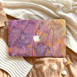 Puede incluir: Una funda rígida para portátil con un diseño abstracto de remolinos morados y naranjas, detalles brillantes y motivos en forma de estrella. El centro de la funda muestra un recorte para el logotipo de Apple plateado, creando un aspecto vibrante.