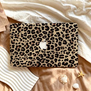 May include: A rectangular laptop cover featuring a tan background with a repeating black and brown leopard print pattern. The cover has a central cutout revealing the white Apple logo. The design covers the entire surface of the laptop lid.
