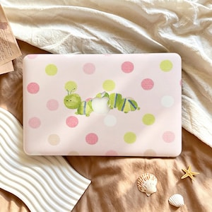 May include: A light pink laptop cover featuring a repeating pattern of soft green, white, and pink polka dots. A watercolor-style green caterpillar illustration is positioned across the center, incorporating the silver Apple logo cutout.