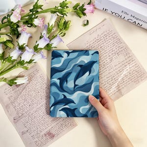 May include: A tablet case with a blue and white dolphin pattern. The case is held by a hand and is surrounded by flowers and vintage paper.