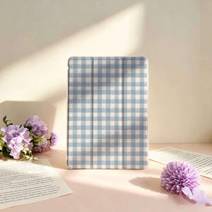 Può includere: Una custodia rettangolare per tablet con un motivo a quadretti vichy azzurro e bianco. La custodia presenta un design a tre ante con pieghe verticali visibili sulla superficie anteriore, conferendo un aspetto strutturato alla protezione.