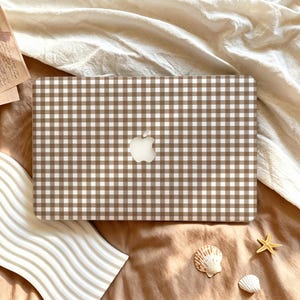 May include: A rectangular laptop cover featuring a brown and white gingham check pattern. The cover has a centered cutout revealing the silver Apple logo. The design consists of a repeating grid of small squares in alternating brown and white colors.