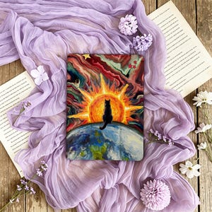 Puede incluir: Una funda rectangular para tableta con una vibrante ilustración de un gato negro sentado sobre la Tierra, mirando hacia un gran sol radiante con nubes arremolinadas en rojo, naranja y amarillo, acentuado por pequeñas estrellas.