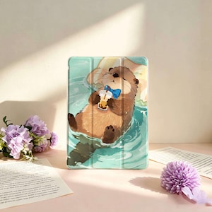 May include: A rectangular tablet case featuring a printed illustration of a brown otter floating in teal water. The otter wears a blue bow tie and holds a plastic cup with a straw containing a tan beverage. The case has a smooth, glossy finish.