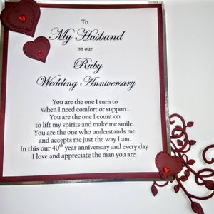 May include: A white card with a red border and silver trim. The card has a message that reads "To My Husband on our Ruby Wedding Anniversary." The message continues with a heartfelt sentiment about love and appreciation. The card is decorated with red hearts.