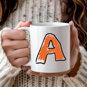 May include: White ceramic mug featuring a large, orange letter "A" with a black outline. The letter has a hand-drawn appearance. The mug is held by a person wearing a white sweater.