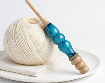 May include: A wooden crochet hook with a teal and wood handle rests on a white plate next to a ball of cream-colored yarn and a folded white cloth. The hook is marked with "6.0mm".
