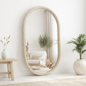 May include: A large oval floor mirror featuring a thick, textured beige rope frame. The mirror has a reflective glass surface that shows a room interior with a white armchair, a small wooden side table, and a potted green plant.
