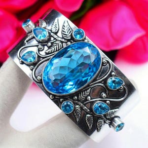 May include: A wide silver cuff bracelet featuring a large, central oval-cut blue topaz gemstone surrounded by smaller, faceted blue topaz stones in pear and round shapes. The silver surface is decorated with intricate, raised leaf and vine motifs.