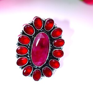 May include: A silver ring featuring a large, oval-shaped, faceted ruby center stone, surrounded by a halo of smaller, round, red gemstones. The ring has a detailed, ornate setting.