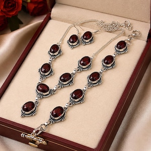 Peut inclure: Un ensemble de bijoux composé d'un collier, d'un bracelet et de deux boucles d'oreilles pendantes en argent. Chaque pièce présente des pierres facettées rouge foncé de forme ovale serties dans des montures en métal argenté ouvragé.