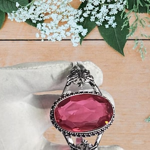May include: A silver-toned bracelet featuring an oval, pink gemstone. The gemstone is surrounded by a black beaded border and set in a decorative, openwork frame. The bracelet is held by a gloved hand, with a wooden background and floral accents.