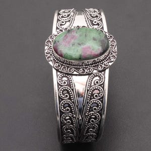 May include: Silver cuff bracelet with an oval gemstone in shades of green and red. The bracelet has an ornate, engraved design with intricate patterns. The hinged design allows for easy wear.