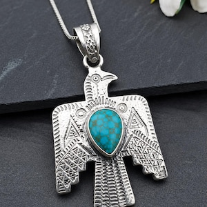 May include: A silver-toned metal pendant shaped like a stylized bird with outstretched wings, featuring a central teardrop-shaped turquoise stone with brown matrix patterns. The pendant is suspended from a thin silver-toned chain necklace.