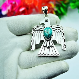 May include: Silver-tone pendant in the shape of a stylized bird, with a turquoise-coloured stone at the centre. The pendant has detailed etched patterns on the wings and tail. The pendant is held in a gloved hand.