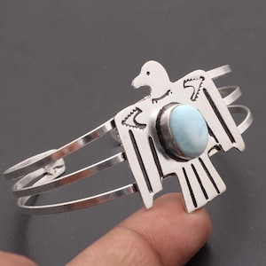 May include: Silver cuff bracelet featuring a stylized bird design with a central oval, light blue gemstone. The bracelet has three parallel bands and a decorative bird element.