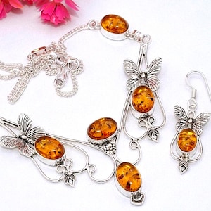 Può includere: Un set di gioielli composto da una collana e un paio di orecchini pendenti. La collana presenta motivi a farfalla in metallo argentato e cabochon di ambra ovali. Gli orecchini abbinati mostrano anch'essi un design a farfalla con pietre d'ambra.