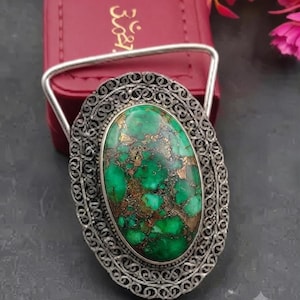 May include: An oval belt buckle featuring a large, polished green stone with brown matrix inclusions. The stone is set within an intricate, dark silver-toned filigree frame that has a decorative, open-work pattern surrounding the central gemstone.