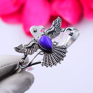 Pode incluir: Uma pulseira prateada com um design de pássaro, incorporando uma pedra preciosa roxa em forma de gota. As asas e a cauda do pássaro são finamente detalhadas. A pulseira está sobre um fundo rosa e branco.