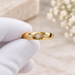 Può includere: Un anello a fascia in oro con tre incastonature a forma di stella, ciascuna contenente una piccola pietra preziosa rotonda e trasparente. La stella centrale è più grande delle due laterali. La superficie dell'anello è liscia e lucida.
