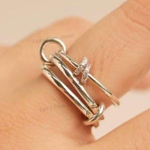 May include: A silver ring with a unique design. The ring features three parallel bars connected by a loop and a knot, with two smaller rings adorned with small, clear stones. The text "Fairy Fine Jewel" is visible.