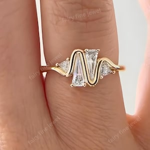 Peut inclure: Une bague en or au design unique, ornée de trois pierres blanches. La pierre centrale est rectangulaire, flanquée de deux pierres triangulaires. La monture de la bague présente un motif ondulé. La bague est portée au doigt.