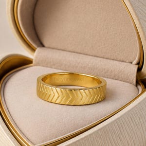 Puede incluir: Un anillo de banda en tono dorado con un patrón repetitivo de chevrón o espiga grabado alrededor de la superficie exterior. El anillo está colocado dentro de un joyero en forma de corazón con un forro interior beige claro y el texto fairyfinejewel.