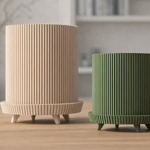 Könnte beinhalten: Zwei zylindrische Pflanzgefäße mit vertikalen Rippendetails. Eines ist hellbeige und das andere ist grün. Beide Pflanzgefäße haben einen passenden Sockel mit drei kleinen Beinen. Diese dekorativen Pflanzgefäße eignen sich für Zimmerpflanzen.