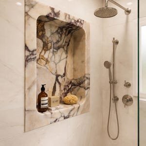 May include: A marble shower niche with a brown soap dispenser, a bar of soap, and a natural sponge. The niche is framed by a marble surround with purple veining. A shower head and handheld shower are visible.