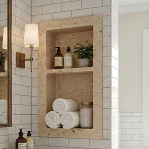 May include: A bathroom with a wooden vanity, white countertop, and a white rectangular sink. The walls are tiled with white subway tiles. A built-in shelf holds rolled white towels, plants, and toiletries. A gold-framed mirror and sconce add elegance.