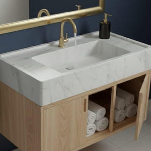 May include: A modern bathroom vanity with a white marble countertop and a light wood cabinet. The vanity has a built-in sink with a gold faucet, a gold-framed mirror, and a towel rack. Rolled white towels are stored in the open cabinet.