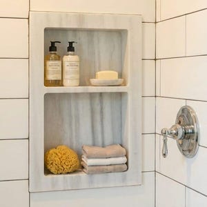 May include: A built-in marble shower niche with two pump bottles, a bar of soap, a natural sponge, and folded towels. The shower is tiled with white rectangular tiles and features a chrome shower head and faucet.