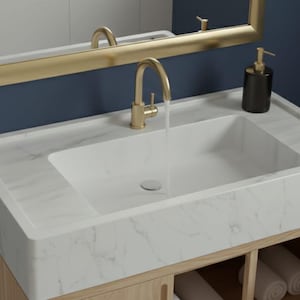 May include: A modern bathroom vanity with a white marble countertop and a rectangular sink. The faucet and mirror frame are gold. A black soap dispenser sits on the counter. A white towel hangs on a gold towel rack. The vanity is made of light wood.