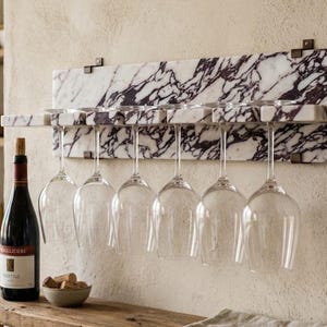 May include: A wall-mounted wine glass rack made of white marble with dark purple veining. The rectangular rack features six slots for hanging stemware, with six clear glass wine glasses suspended upside down from the marble base.