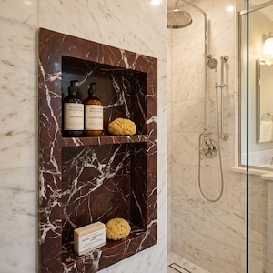 May include: A built-in shower niche with a dark red marble finish, holding shampoo, conditioner, a bar of soap, and a natural sponge. The shower features a rainfall showerhead and a glass enclosure, set against a backdrop of white marble tiles.
