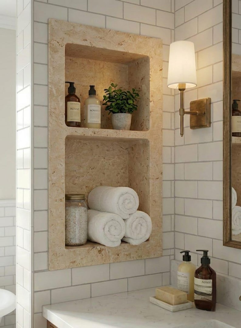 May include: A built-in bathroom shelf with two levels, holding toiletries, a small potted plant, and rolled white towels. The shelf is made of a light beige stone and is set against white subway tile.